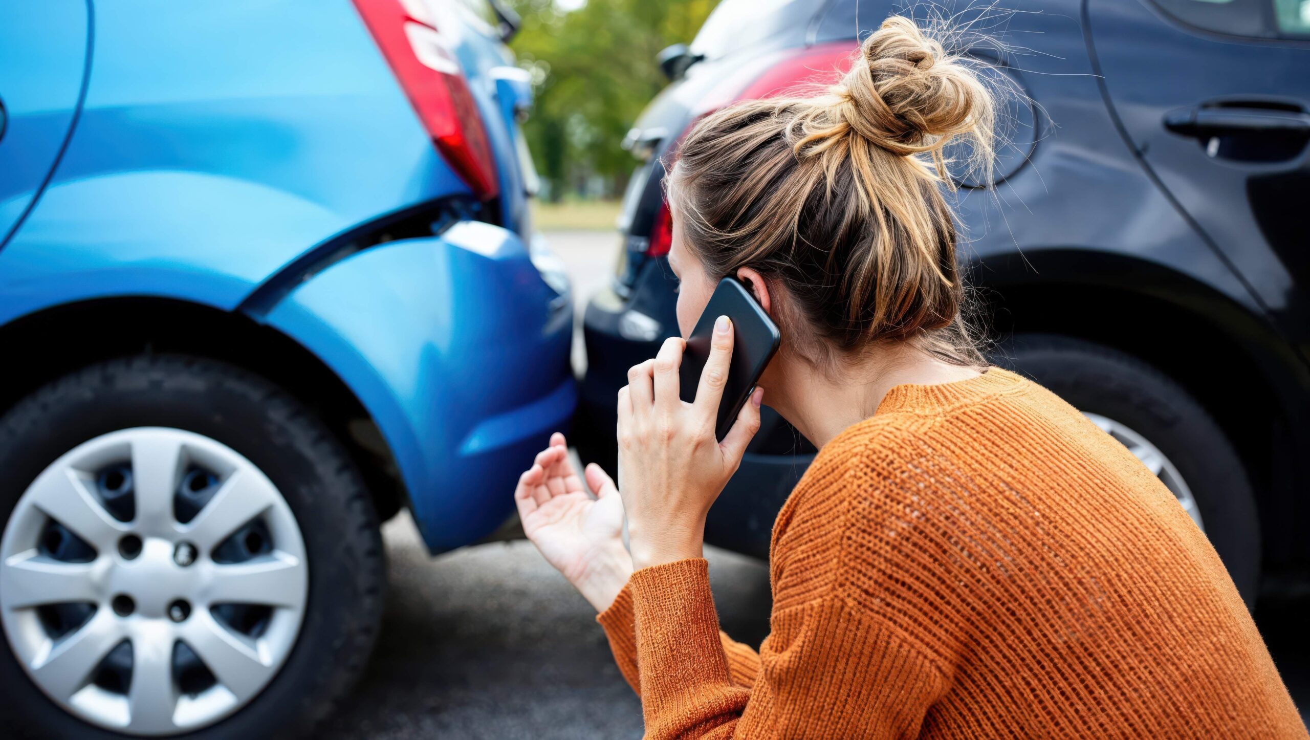Woman in orange sweater calling on phone at scene of car accident between blue and dark vehicles