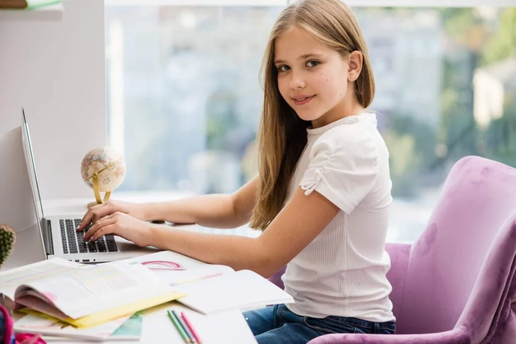 Young girl with long blonde hair sitting at a desk with a laptop, globe, and study materials, smiling while typing.